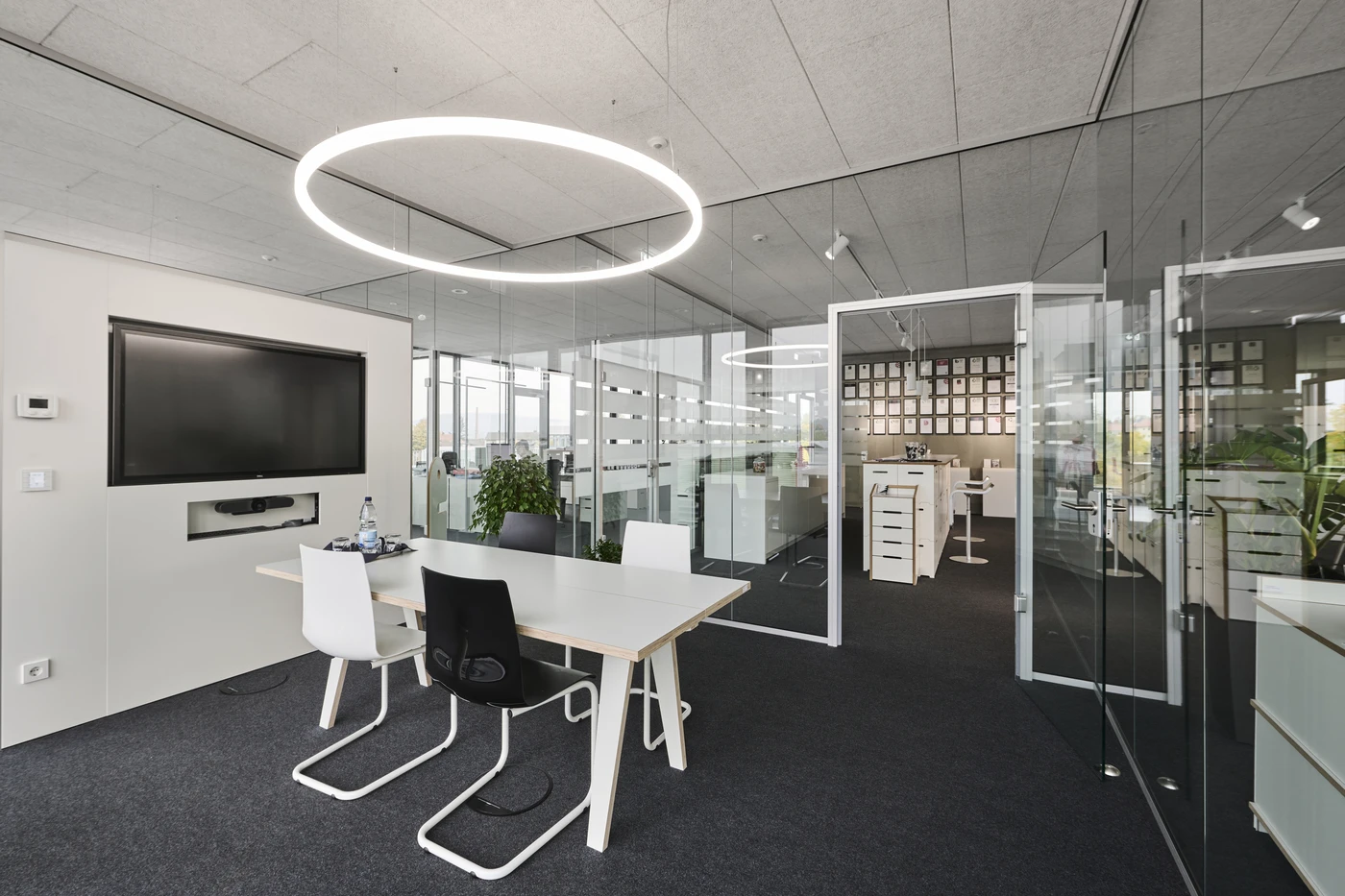 Projekte Ein modernes Büro mit Glaswänden, einem Konferenztisch mit vier Stühlen, einem großen Wandmonitor und einer runden LED-Deckenleuchte. Der Raum ist hell und offen, mit Pflanzen und mehreren Arbeitsplätzen ausgestattet – ideal für die Generalplanung und Projektentwicklung in allen Leistungsphasen.
