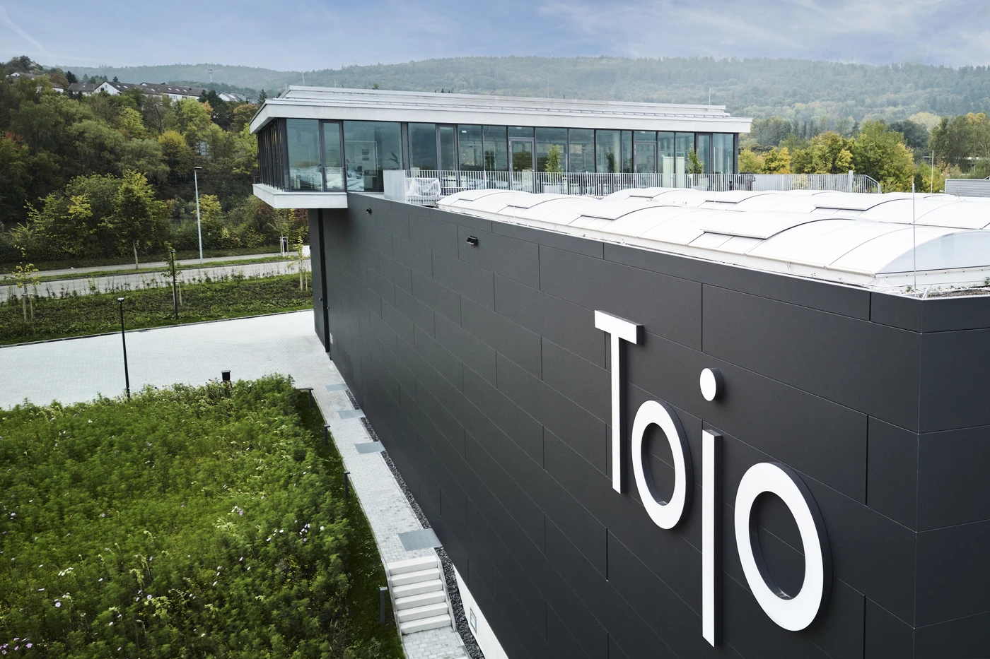 Projekte Modernes Gebäude mit großen weißen „Tojo“-Buchstaben an einer dunklen Außenwand, entworfen von SCHATZ Architektur, umgeben von Grünflächen und Bäumen in Schorndorf, mit einem verglasten Obergeschoss und fernen Hügeln im Hintergrund.