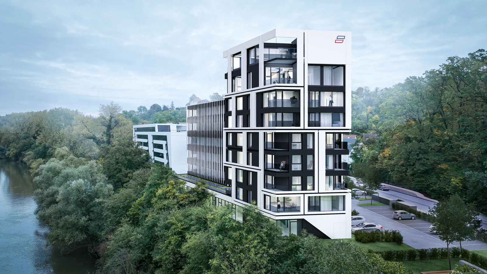 Ein modernes, mehrstöckiges Gebäude mit großen Glasfenstern und Balkonen steht an einem Fluss, umgeben von Bäumen und Grünflächen, mit einer Straße und geparkten Autos in der Nähe.