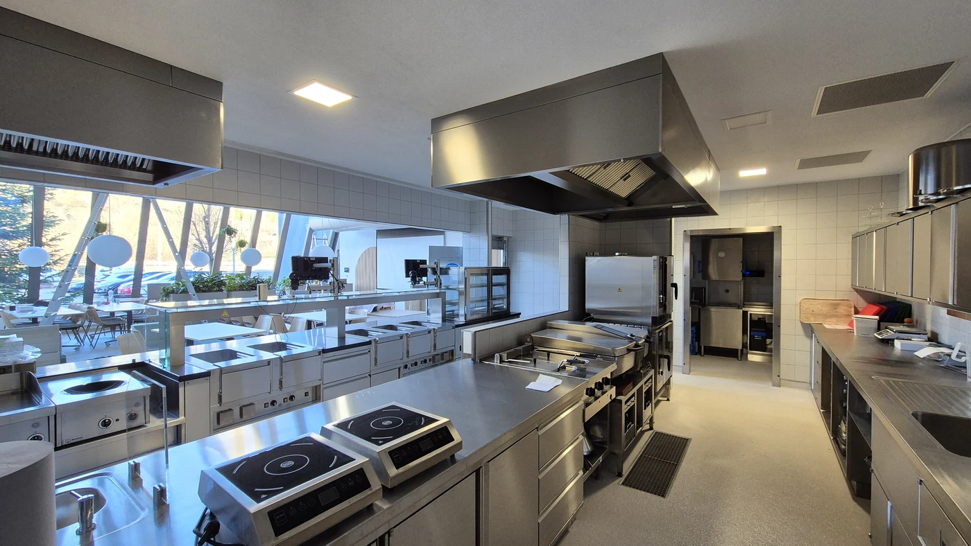 Umbau Betriebsrestaurant Pfisterer Winterbach Moderne Großküche mit Edelstahlgeräten, großen Herden, Backöfen, Arbeitsflächen und einem Fenster mit Blick auf einen Sitzbereich im Freien. Der von SCHATZ Architektur in Schorndorf entworfene Raum wirkt klar, übersichtlich und ist durch Tageslicht und künstliche Beleuchtung gut ausgeleuchtet – ein Zeichen für die sorgfältige Planung in allen Leistungsphasen.