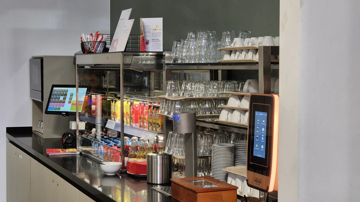 Umbau Betriebsrestaurant Pfisterer Winterbach An einer Selbstbedienungstheke in Schorndorf sind Gläser, Tassen und Saftflaschen gestapelt, außerdem gibt es einen Touchscreen-Getränkeautomaten, eine Kasse, Besteck sowie ausgehängte Speisekarten. Die Theke ist ordentlich und übersichtlich – ein Beweis für eine effiziente Gesamtplanung.