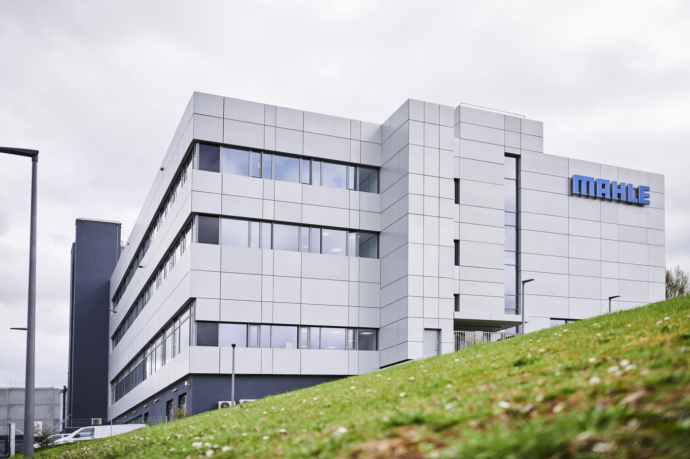 Startseite Ein modernes, graues Bürogebäude mit großen Fenstern und dem MAHLE-Logo in der oberen rechten Ecke, gesehen von einem grasbewachsenen Hügel unter bewölktem Himmel – ein eindrucksvolles Beispiel für Projektentwicklung und Generalplanung über alle Leistungsphasen hinweg.