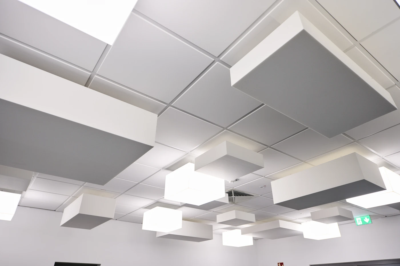 Weiße Decke mit rechteckigen Paneelen und abgehängten quadratischen Leuchten, von denen einige beleuchtet sind. Im Hintergrund ist ein grünes Notausgangsschild zu sehen. Der Entwurf besticht durch ein modernes und geometrisches Design, das eine klare und minimalistische Ästhetik schafft, inspiriert von aktuellen Projekten in Schorndorf.