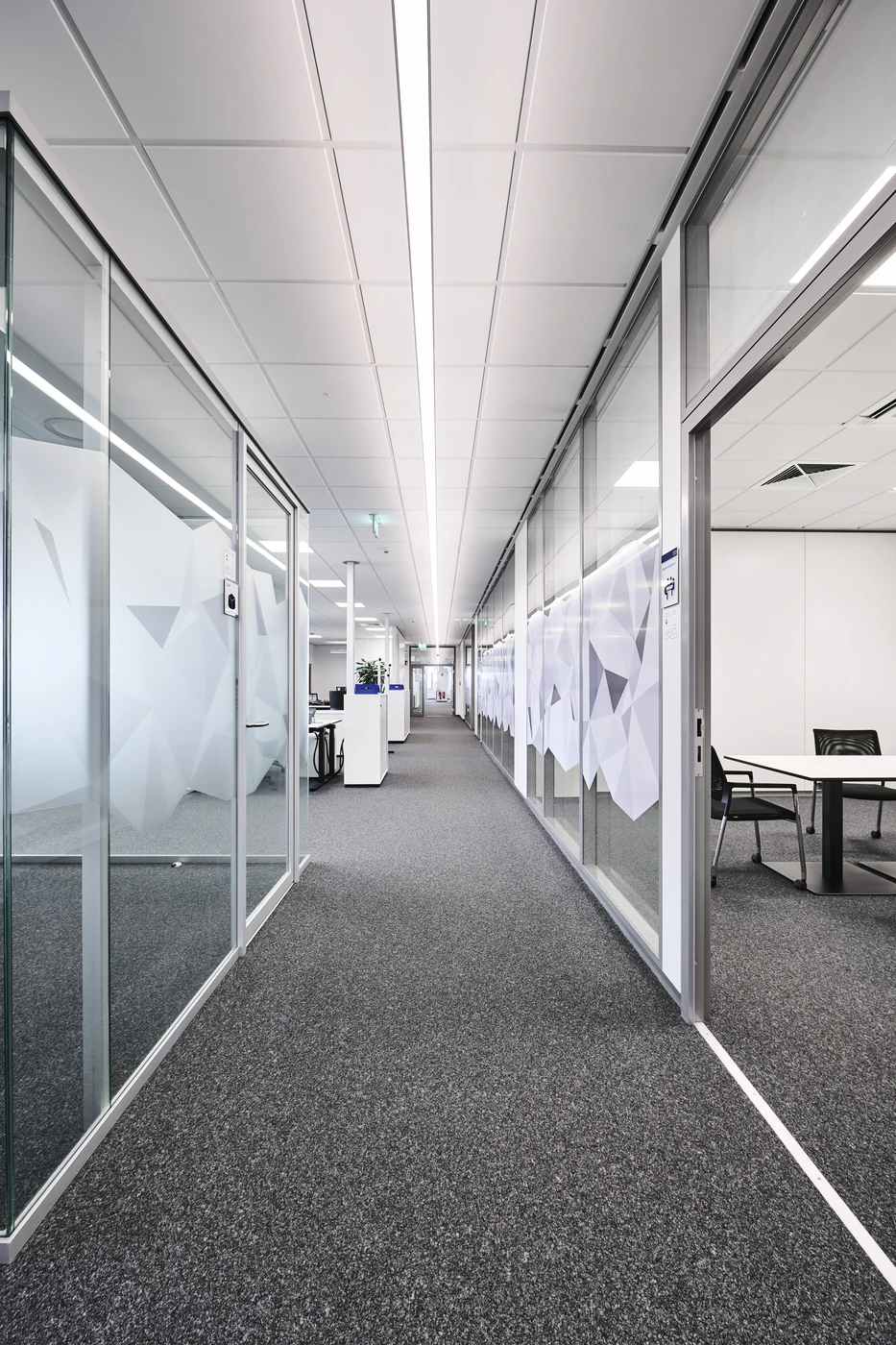 Ein moderner Büroflur mit grauem Teppichboden, beidseitig durch Glaswände abgegrenzten Besprechungsräumen, geometrischen Mattglasmustern an den Scheiben sowie weißen Deckenplatten und heller Deckenbeleuchtung – ein vorbildliches Design von SCHATZ Architektur aus Schorndorf, das über alle Leistungsphasen hinweg gekonnt umgesetzt wurde.