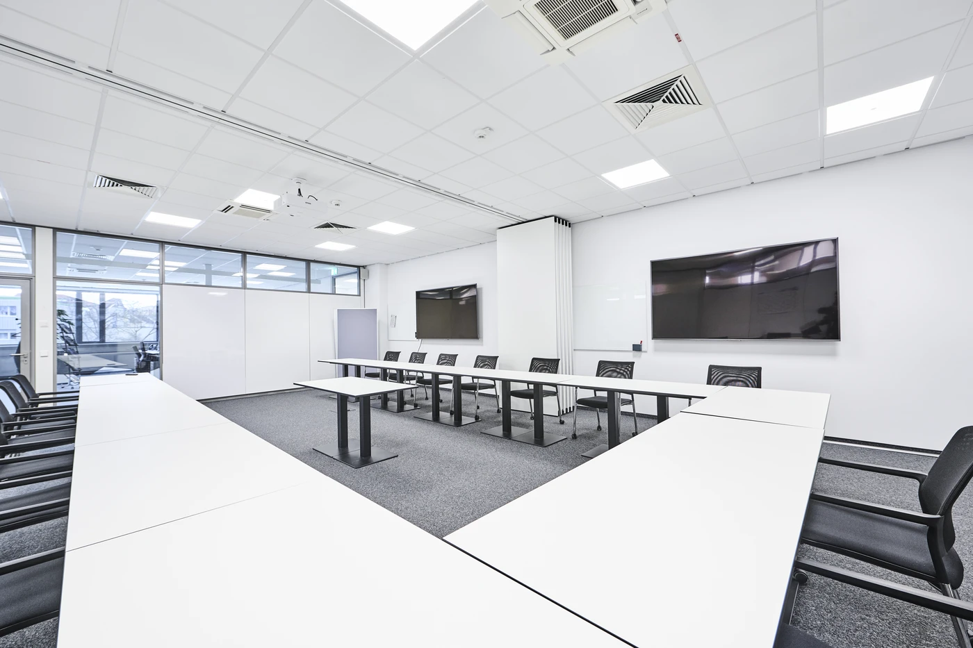 Ein moderner Konferenzraum mit weißen Wänden, weißen Tischen in U-Form, schwarzen Stühlen, zwei großen an der Wand montierten Bildschirmen und Glasfenstern, durch die Tageslicht hereinströmt – dieser helle, minimalistische Raum ist ein Paradebeispiel für die Ergebnisse einer sorgfältigen Projektentwicklung und Generalplanung. Deckenleuchten und Teppichboden runden das durchdachte Raumkonzept ab.