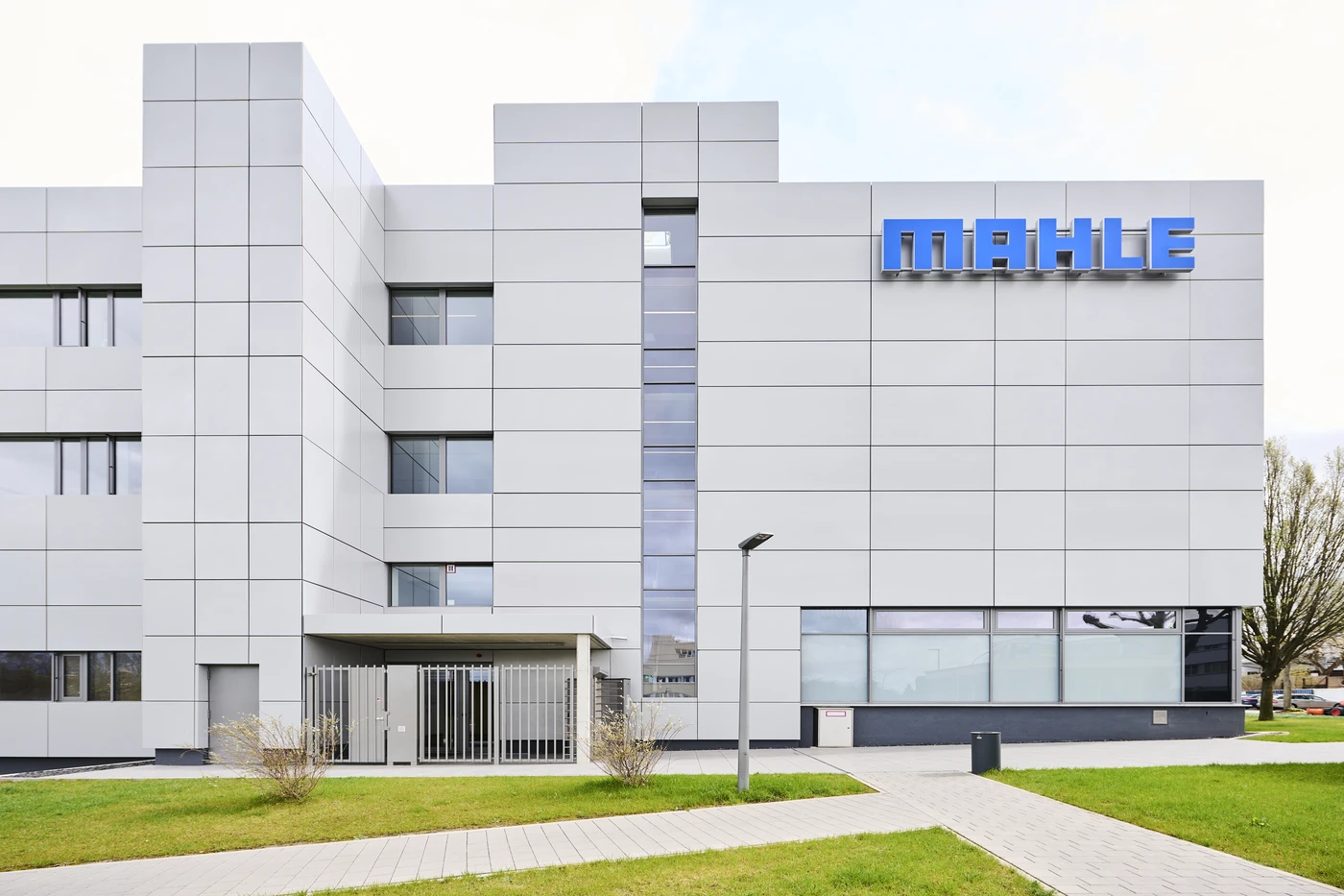 Ein modernes, graues Gebäude mit großen Fenstern und dem blauen „MAHLE“-Logo oben rechts präsentiert den innovativen Entwurf von SCHATZ Architektur. Davor befinden sich ein Tor, ein Weg, Rasen und einige kleine Sträucher.