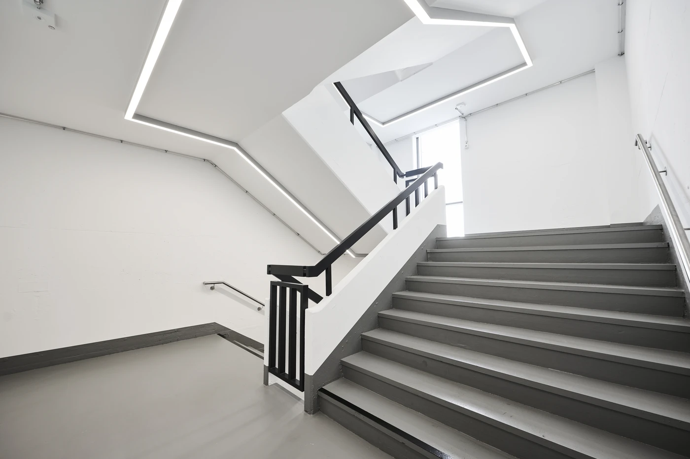 Projekte Ein modernes, minimalistisches Treppenhaus mit grauen Stufen, weißen Wänden, schwarzen Handläufen und geometrischen LED-Deckenleuchten – ein vorbildlicher Entwurf, der durch eine durchdachte Projektentwicklung umgesetzt wurde; durch ein großes Fenster strömt Tageslicht herein.