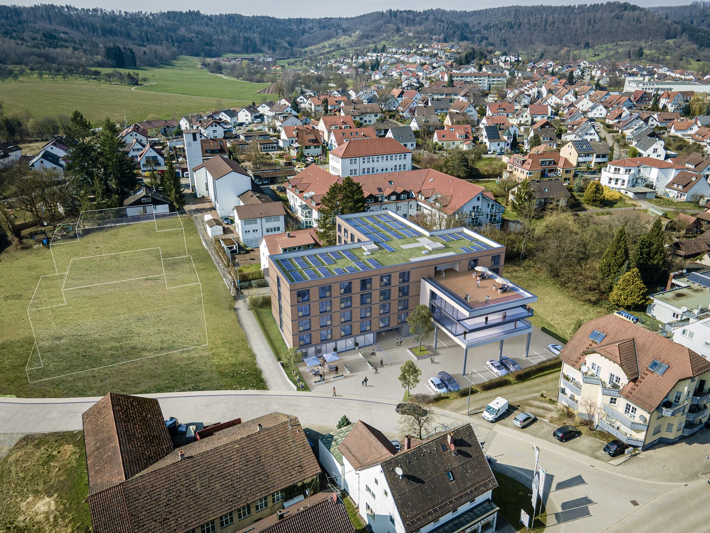 Luftaufnahme von Schorndorf mit einem modernen Gebäude mit Solaranlage, das im Rahmen der Generalplanung von SCHATZ Architektur entworfen wurde und von Häusern, grünen Feldern und einem Sportgelände mit kaum erkennbaren Spielfeldmarkierungen umgeben ist. Im Hintergrund sind bewaldete Hügel zu sehen.