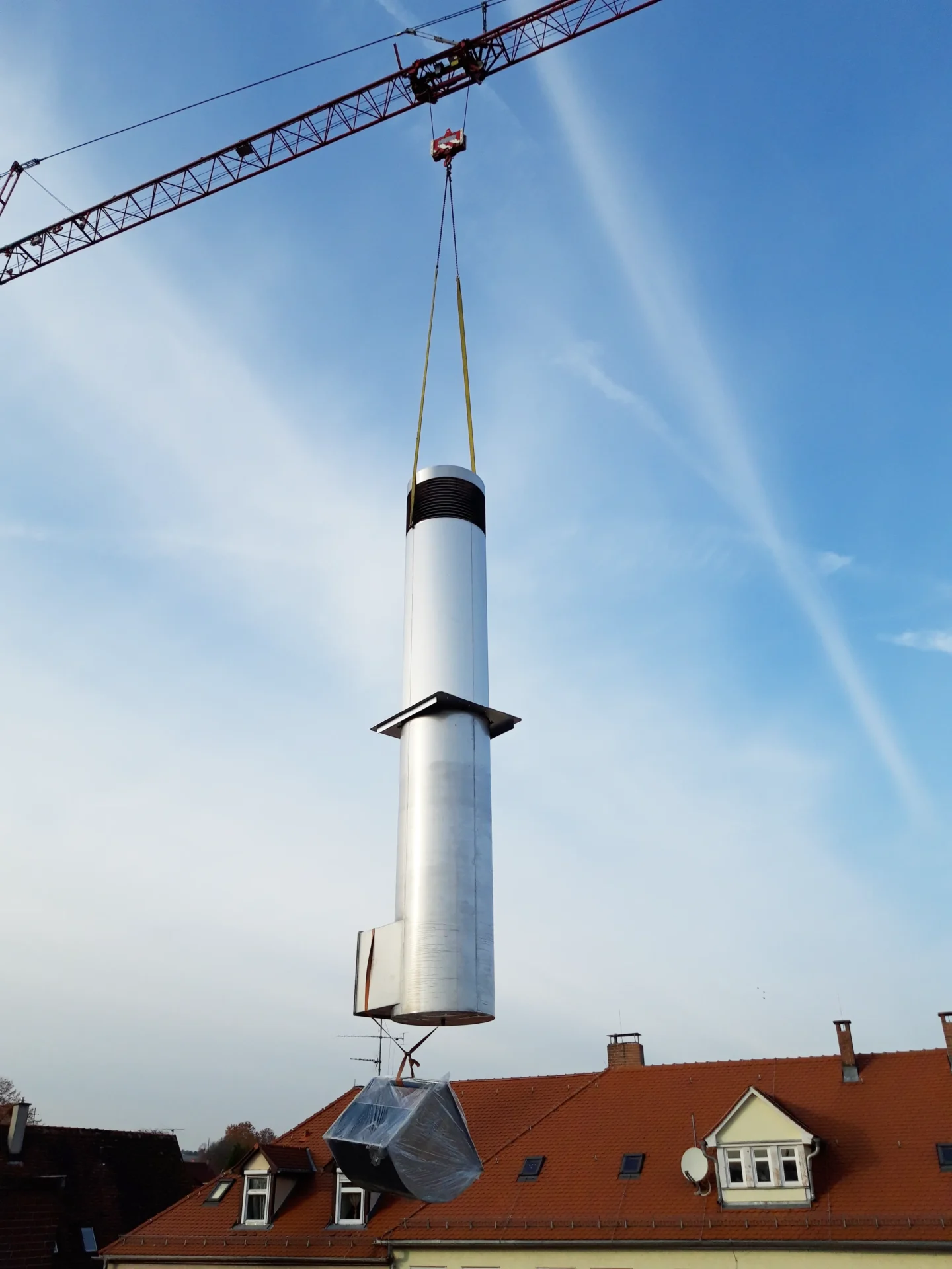 Umbau Pflegestift nach LHeimBauVO, Stuttgart-Münster Ein Kran hebt einen großen, zylinderförmigen Metallkörper, der einer Rakete ähnelt, über Gebäude mit roten Dächern hinweg, vor einem blauen Himmel mit vereinzelten Wolken.