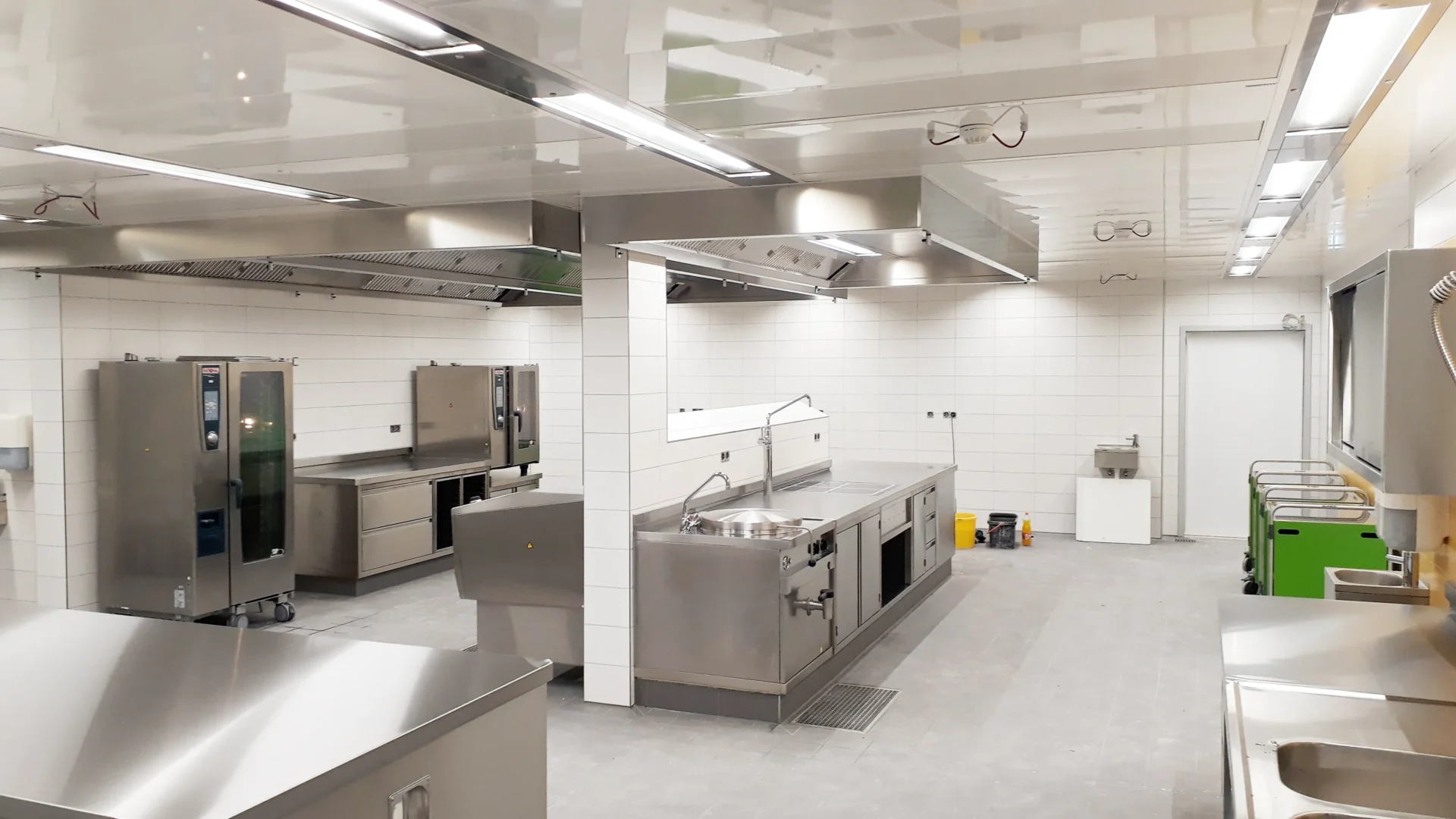 Umbau Pflegestift nach LHeimBauVO, Stuttgart-Münster Eine geräumige, moderne Großküche mit Edelstahlgeräten, weiß gefliesten Wänden und grauen Fußböden. Zu sehen sind mehrere Arbeitsstationen, Öfen und Spülbecken, die von heller Deckenbeleuchtung erhellt werden und einen aufgeräumten, sauberen Eindruck machen.