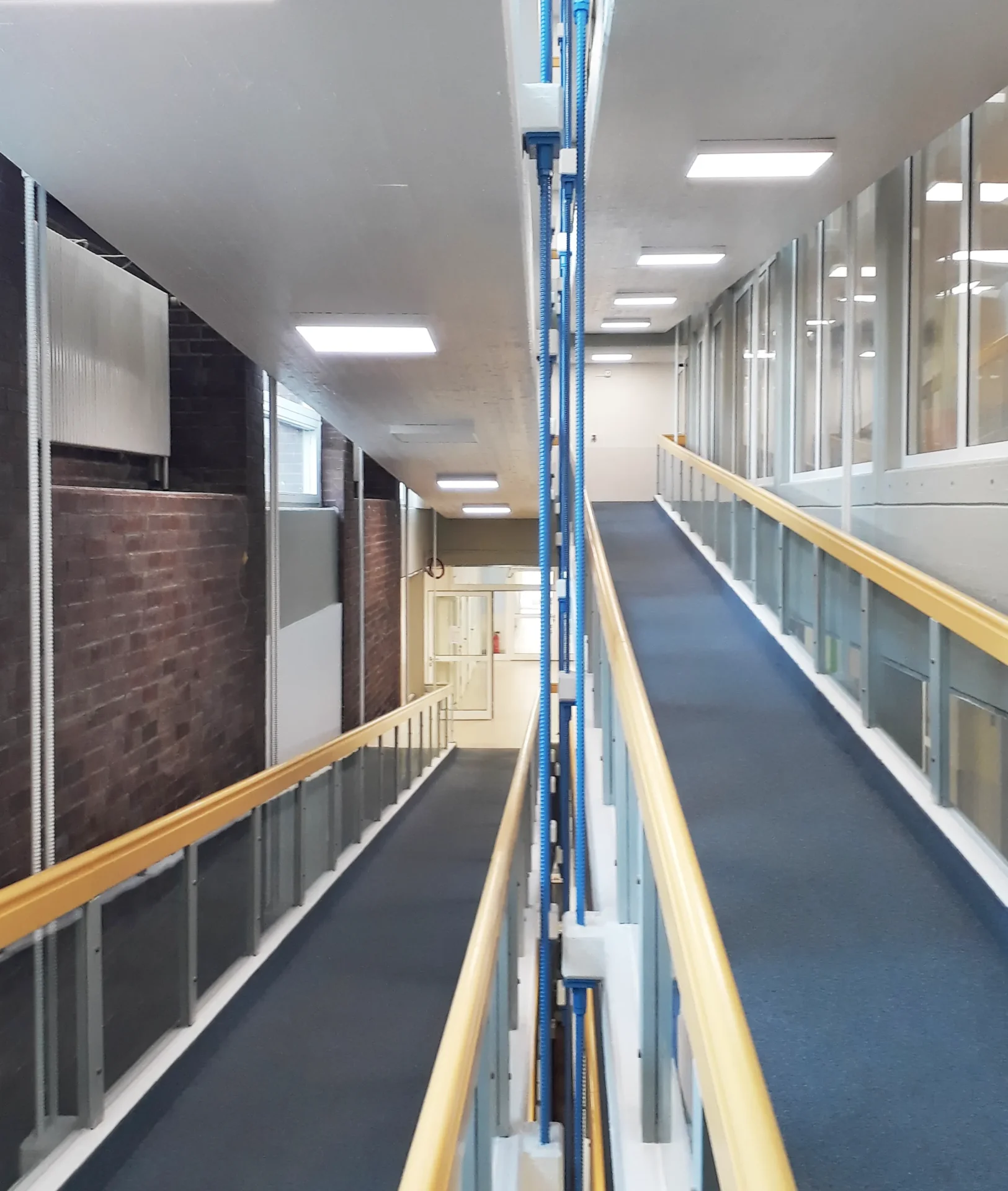 Umbau Pflegestift nach LHeimBauVO, Stuttgart-Münster Zwei mit blauem Teppich ausgelegte, barrierefreie Rampen mit gelben Handläufen verlaufen parallel zwischen Backstein- und Glaswänden in einem hell beleuchteten Flur und führen zu einer Tür am anderen Ende.