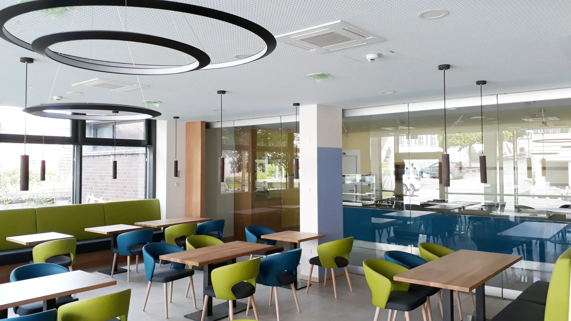 Umbau Pflegestift nach LHeimBauVO, Stuttgart-Münster Moderne Cafeteria mit Holztischen, blauen und grünen Stühlen sowie großen runden Deckenleuchten. Es gibt Glastrennwände, eine Ausgabetheke und große Fenster, durch die Tageslicht hereinfällt.