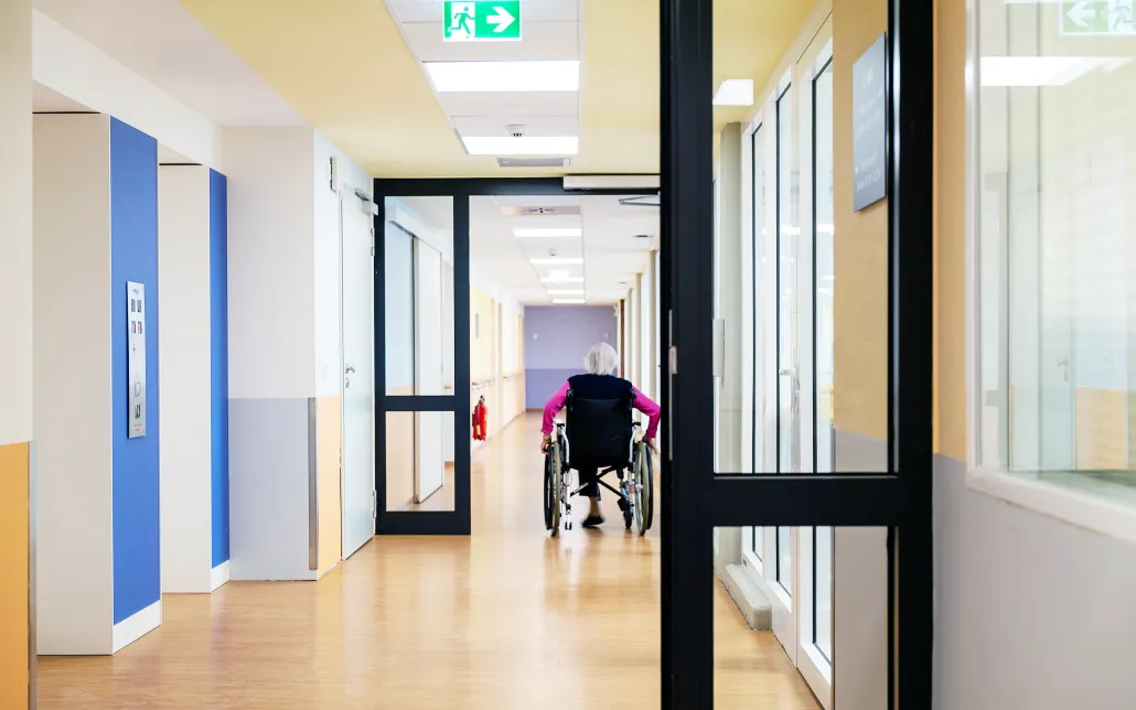 Umbau Pflegestift nach LHeimBauVO, Stuttgart-Münster Eine ältere Person im Rollstuhl fährt einen hell beleuchteten, leeren Flur in einer Gesundheitseinrichtung oder einem Pflegeheim entlang, an dessen Seiten sich Türen und Fenster befinden.