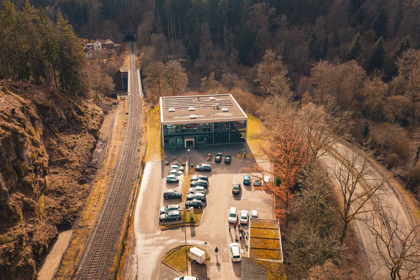 Luftaufnahme eines modernen Gebäudes in der Nähe einer Bahnstrecke in Schorndorf, mit geparkten Autos und Menschen vor dem Gebäude, umgeben von Bäumen und einer kurvenreichen Straße in einem Waldgebiet – ein eindrucksvolles Beispiel für durchdachte Generalplanung und innovative Projektentwicklung.