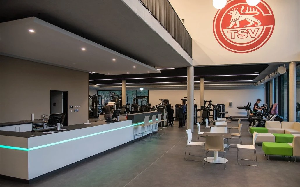 Moderne Fitnessstudio-Innenausstattung mit Empfangstheke, Tischen und Stühlen im Vordergrund, gelegen in Schorndorf. Im Hintergrund nutzen Menschen die Trainingsgeräte unter einem großen roten TSV-Logo an der Wand darüber. Diese klare, großzügige und gut beleuchtete Umgebung spiegelt die Liebe zum Detail in allen Leistungsphasen der Projektentwicklung wider.