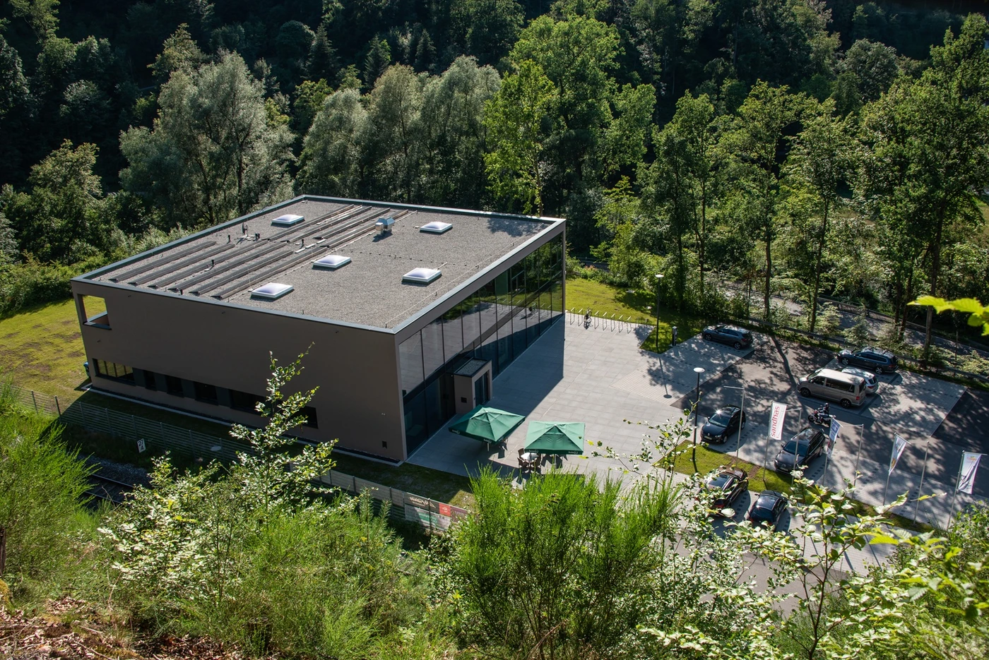 Sport Ein modernes, rechteckiges Gebäude mit Flachdach steht in einer waldreichen Umgebung und verkörpert den durchdachten Entwurf, der für SCHATZ Architektur charakteristisch ist. Auf dem angrenzenden Parkplatz stehen mehrere Autos, und zwei grüne Sonnenschirme spenden den Tischen auf der Terrasse vor dem Gebäude Schatten.