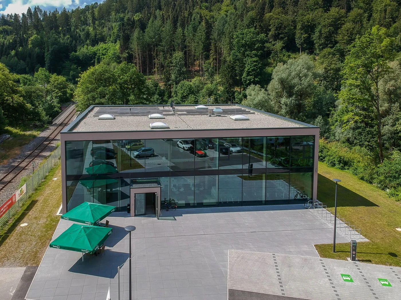 Ein modernes Glasgebäude mit Flachdach liegt in der Nähe eines Waldes und von Bahngleisen. Im Rahmen einer innovativen Projektentwicklung stehen draußen mehrere Autos, und grüne Sonnenschirme spenden den Sitzgelegenheiten am Eingang Schatten. Bäume und Grünflächen umgeben das Gelände.