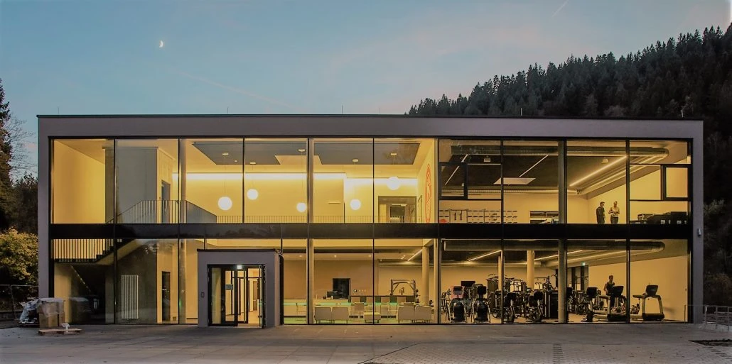 Ein modernes zweistöckiges Gebäude mit großen Glasfenstern und einem hell erleuchteten Innenraum, in dem Fitnessgeräte und einige Personen zu sehen sind; das von SCHATZ Architektur im Rahmen ihres „Generalplanung“-Konzepts entworfene Bauwerk fügt sich in der Abenddämmerung nahtlos in die umliegenden Bäume und Hügel ein.