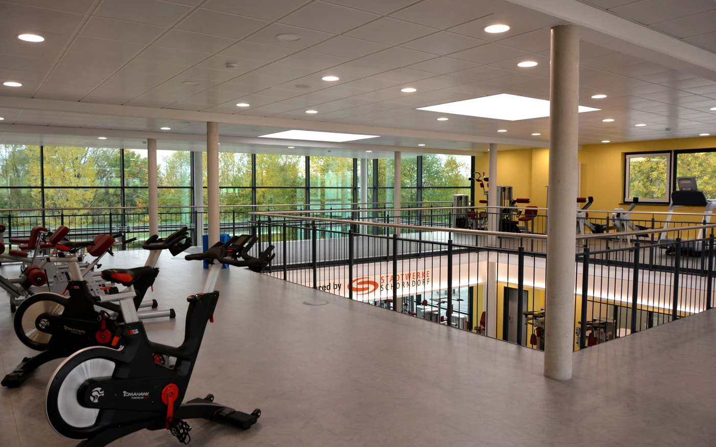 Ein modernes Fitnessstudio in Schorndorf mit Heimtrainern und anderen Fitnessgeräten auf einer oberen Zwischenebene, großen Fenstern und gelben Wänden mit Blick auf eine grüne Außenlandschaft – ein perfektes Beispiel für durchdachte Generalplanung in zeitgenössischen Fitnessräumen.
