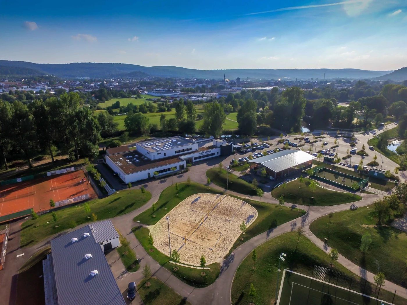 Sport Luftaufnahme eines Sportkomplexes mit Tennisplätzen, einem Sandvolleyballfeld, Parkplätzen, Gebäuden, Bäumen und fernen Hügeln unter strahlend blauem Himmel – ein Beweis für die sorgfältige Generalplanung und den innovativen Entwurf von SCHATZ Architektur.