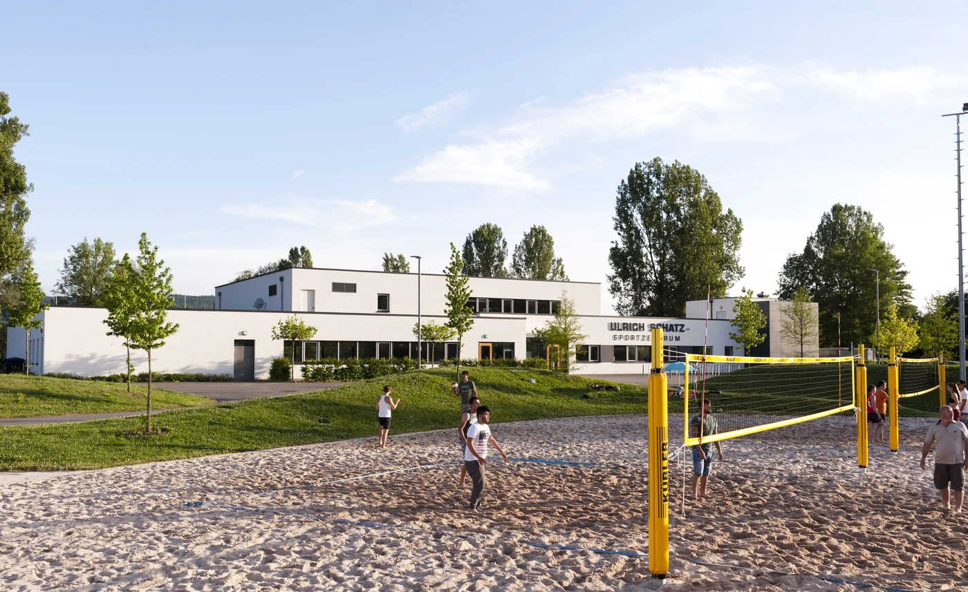 Auf einem sandigen Platz mit gelben Netzen, in der Nähe des modernen, weißen Ulrich-Schütz-Sportzentrums – einem beeindruckenden Projekt von SCHATZ Architektur – spielen die Menschen Beachvolleyball. Umgeben von Bäumen und grünem Rasen in Schorndorf bietet die Anlage eine perfekte Mischung aus Sport und Freizeit unter freiem Himmel.