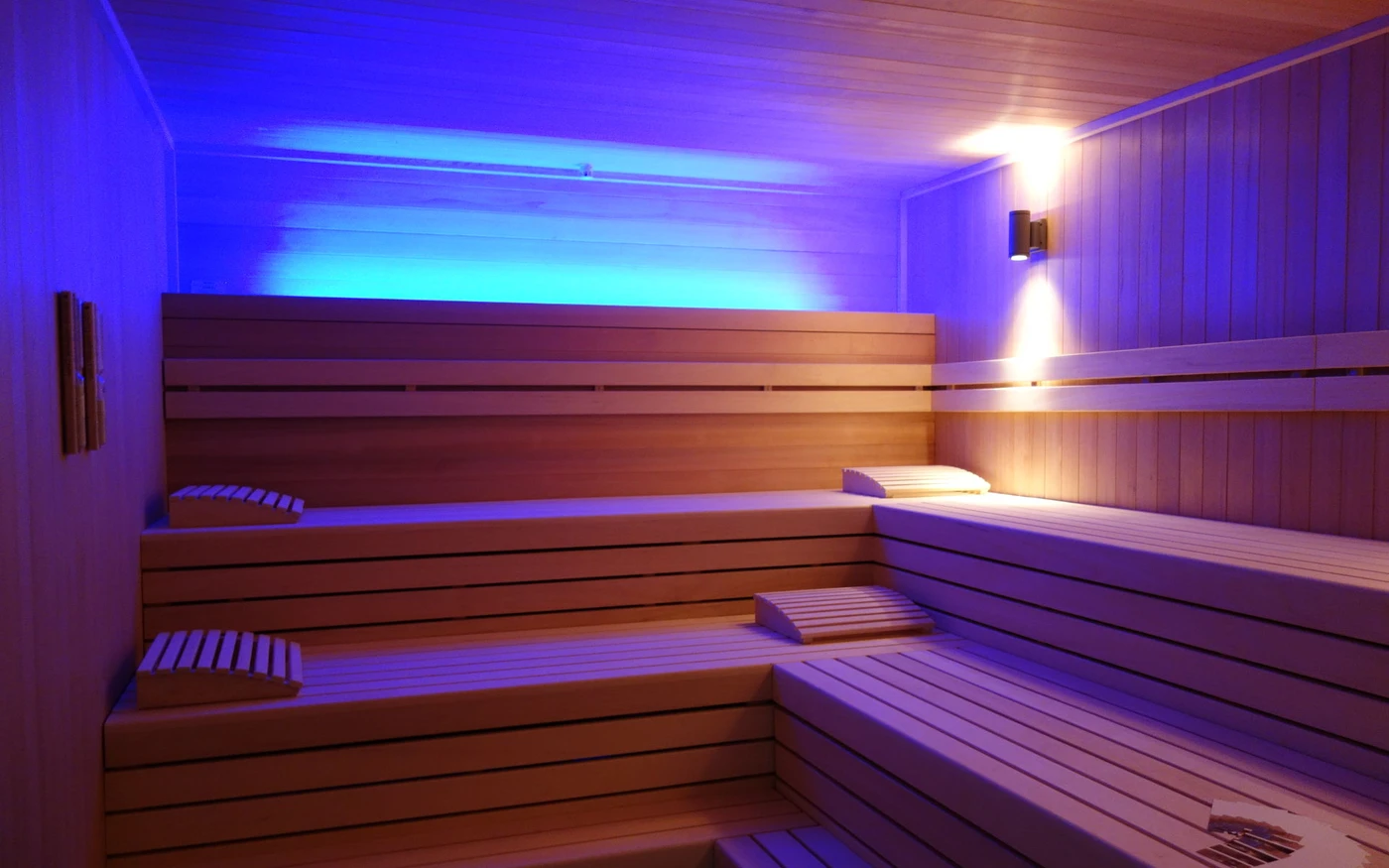 Ulrich Schatz Sportzentrum im Sportpark Rems Das Interieur der Holzsauna mit gestuften Bänken, Kopfstützen und sanfter Stimmungsbeleuchtung in Blau und Gelb schafft eine warme und entspannende Atmosphäre. Der Raum spiegelt den durchdachten Entwurf von SCHATZ Architektur wider und verdeutlicht deren charakteristische Designphilosophie. Es sind keine Personen zu sehen.