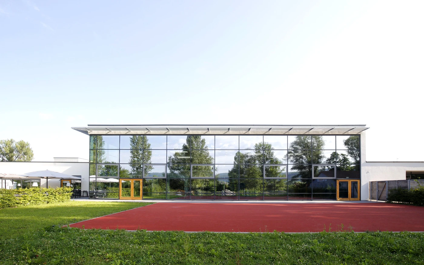 Ulrich Schatz Sportzentrum im Sportpark Rems Ein modernes Gebäude mit großen Glasfenstern, in denen sich Bäume und Himmel spiegeln, das im Rahmen einer Generalplanung in Schorndorf entworfen wurde und sich durch gelb gerahmte Türen sowie einen roten Sportplatz vor dem Haus auszeichnet, der von grünem Gras und Laub umgeben ist.