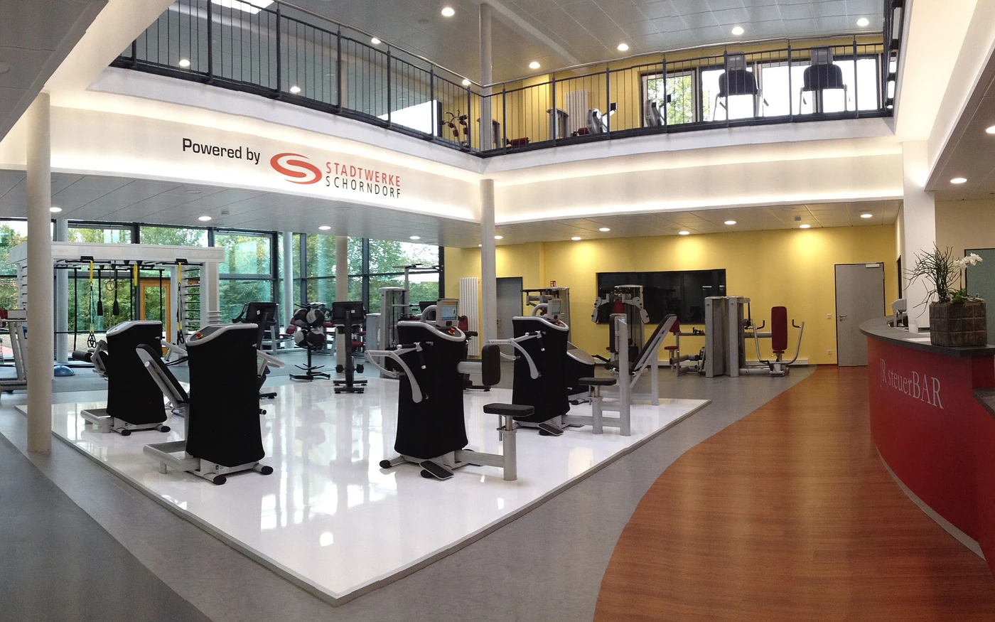 Ulrich Schatz Sportzentrum im Sportpark Rems Modernes Fitnessstudio mit verschiedenen Trainingsgeräten, großen Fenstern und einem Obergeschoss mit weiteren Geräten. Die Wände sind gelb und weiß, und an der Wand hängt ein Schild mit der Aufschrift „Powered by Stadtwerke Schongau“. Das Studio wurde in Zusammenarbeit mit SCHATZ Architektur in der Entwurfsphase gestaltet und ist mit einigen Pflanzen dekoriert.