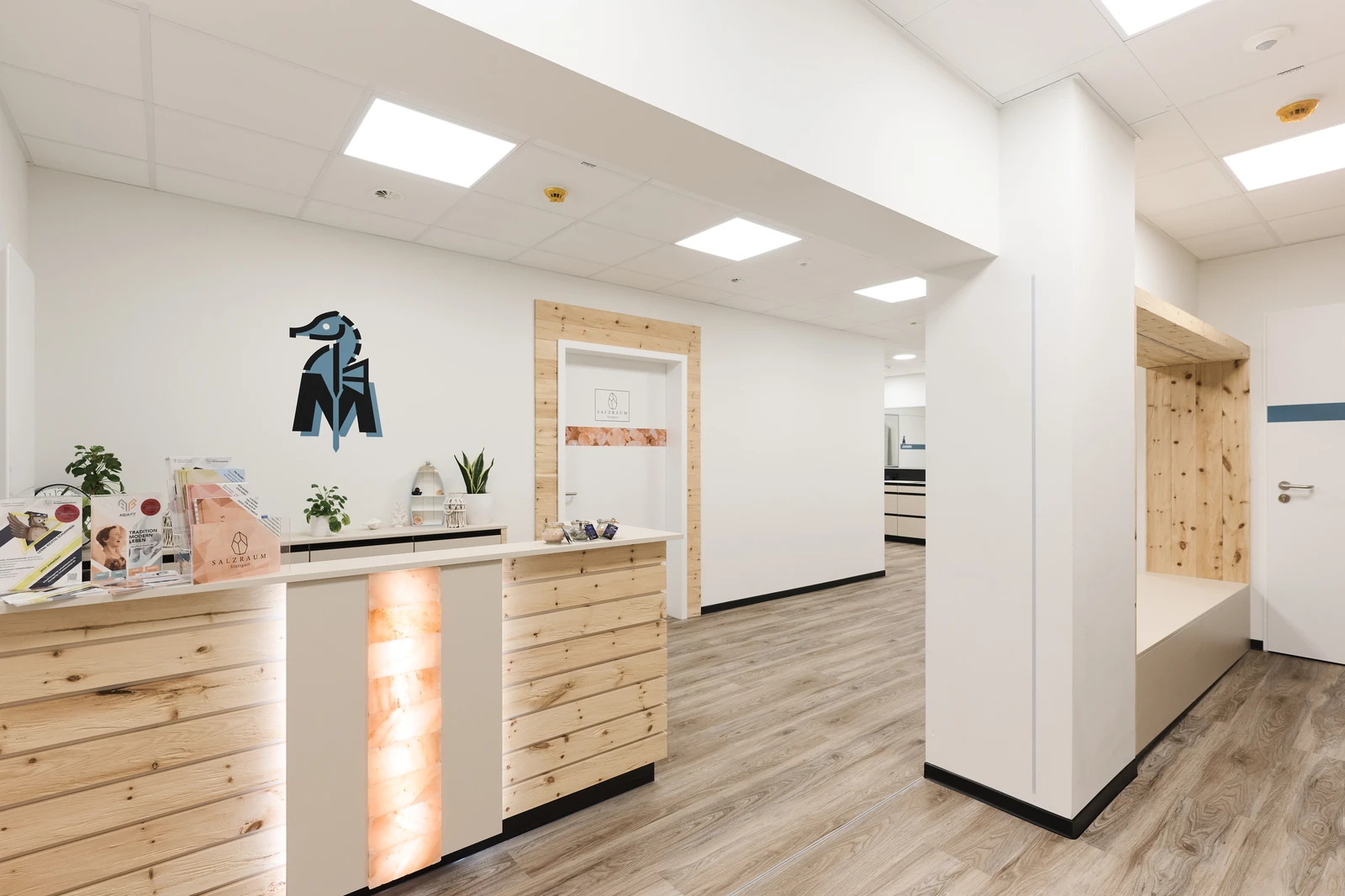 Ein moderner, übersichtlicher Empfangsbereich einer Klinik mit Holzvertäfelung, einer Sitzbank, Broschüren, Pflanzen und einem Seepferdchen-Logo an der Wand. Der Raum ist hell, mit weißen Wänden und einem hellen Holzboden.