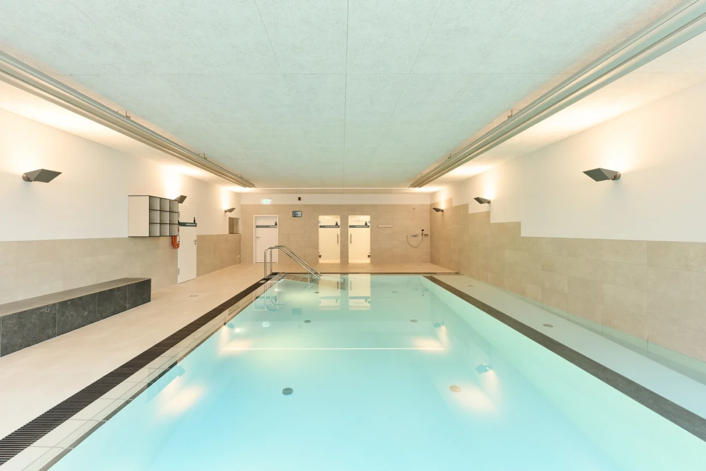 Startseite Helles Hallenbad mit klarem Wasser, beige gefliesten Wänden, Deckenleuchten, einer Bank auf der linken Seite und Türen im Hintergrund. Modernes, minimalistisches Design, keine Personen im Bild.