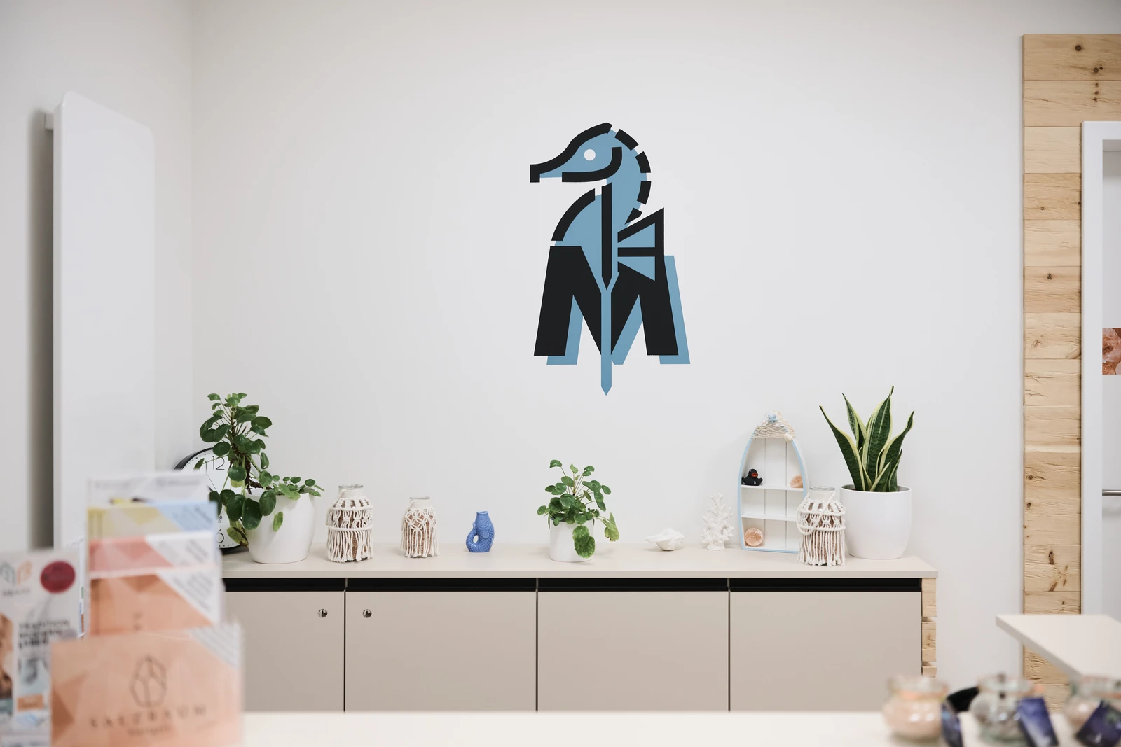 Ein moderner Büroraum mit Pflanzen, Dekoartikeln und einem geometrischen blauen Tierlogo an einer weißen Wand über einem Schrank.
