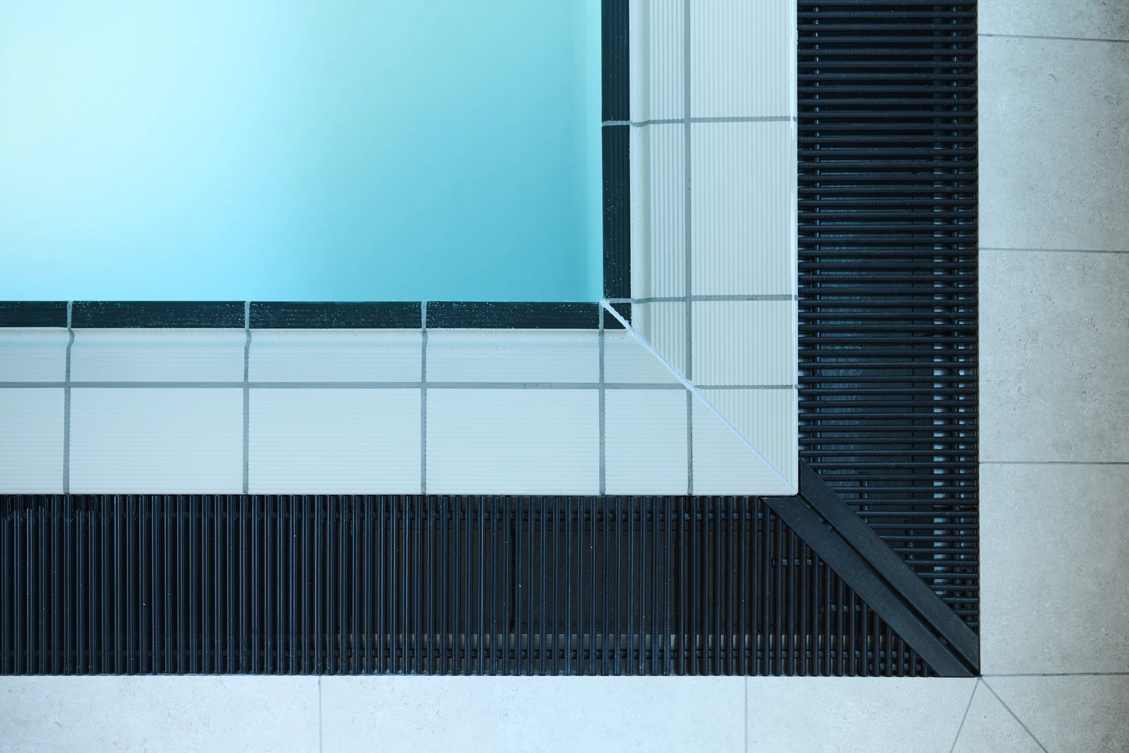 Eine Ansicht von oben auf eine Ecke eines Schwimmbeckens mit blauem Wasser, weißen Fliesen und schwarzen Abflussgittern am Beckenrand.