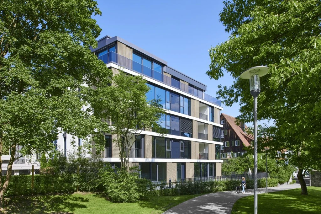 Ein modernes Mehrfamilienhaus mit großen Fenstern, das im Rahmen eines Entwurfs der Schorndorf Projektentwicklung entstanden ist, liegt inmitten von grünen Bäumen und einer Rasenfläche. Ein Gehweg schlängelt sich am Gebäude entlang, und unter strahlend blauem Himmel geht eine Person in der Nähe spazieren.