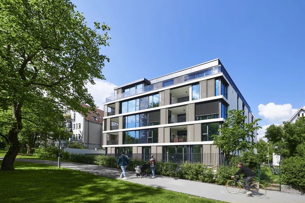 Generalplanung Modernes vierstöckiges Wohnhaus mit großen Glasbalkonen, umgeben von Bäumen in Schorndorf. An einem sonnigen Tag spazieren Menschen, schieben Kinderwagen und fahren mit dem Fahrrad auf einem Weg vor dem Gebäude – eine lebendige Szene, die die durchdachte Generalplanung über alle Leistungsphasen hinweg widerspiegelt.