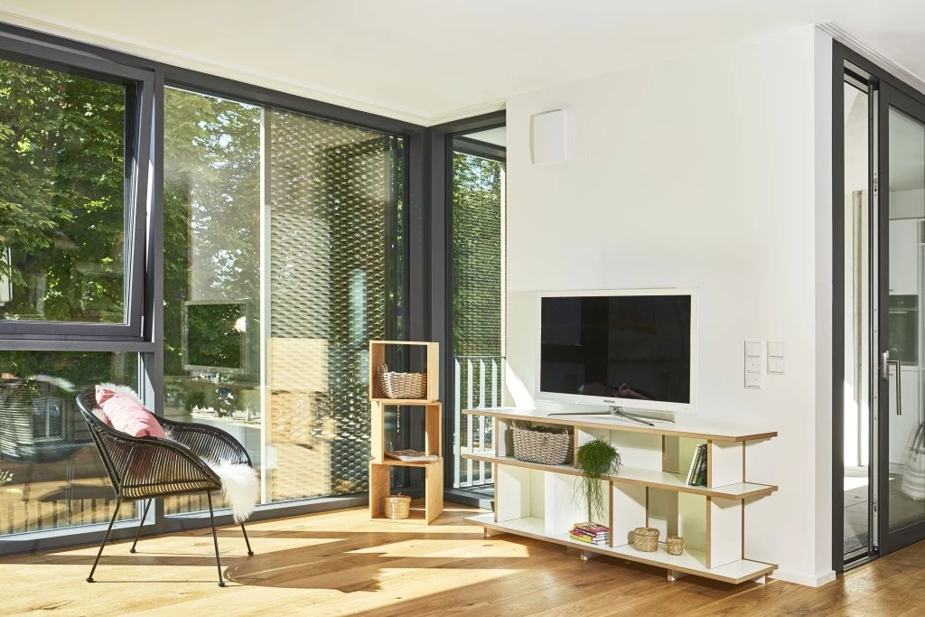Ein helles, modernes Wohnzimmer mit großen Fenstern, Holzboden, einem schwarzen Sessel mit rosa Kissen, kleinen Regalen und einem Fernseher auf einem weißen Ständer; draußen sind grüne Bäume zu sehen – ein perfektes Beispiel für durchdachten Entwurf und Projektentwicklung in der Wohnraumbauplanung.