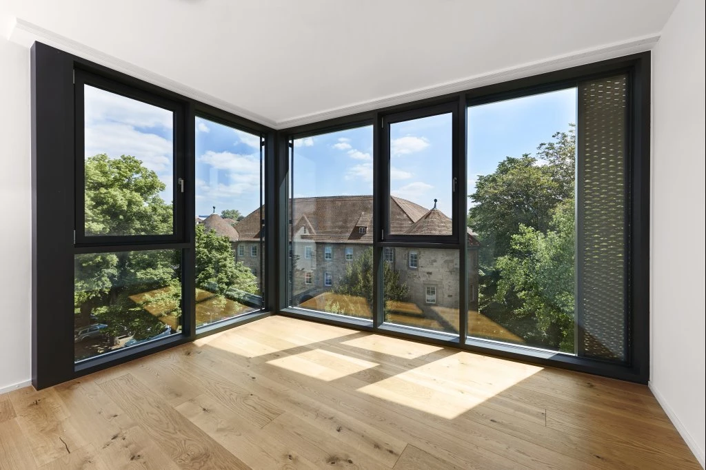 Projekte Ein Eckzimmer mit großen, raumhohen Fenstern, hellem Holzfußboden und einem Blick auf Bäume und ein historisches Gebäude draußen an einem sonnigen Tag – ein einladender Raum, der den durchdachten Entwurf von SCHATZ Architektur über alle Leistungsphasen hinweg widerspiegelt.