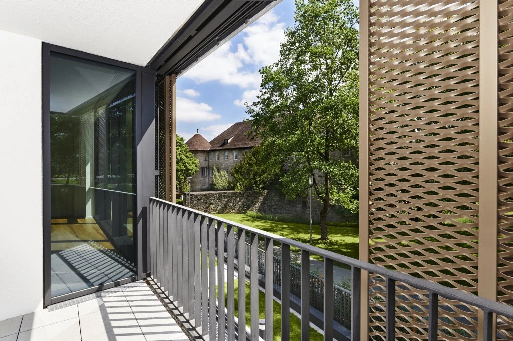Ein moderner Balkon mit Metallgeländern und perforierten Sichtschutzwänden, der in der Entwurfsphase entworfen wurde, bietet unter einem teilweise bewölkten Himmel den Blick auf eine grüne Wiese mit Bäumen und ein altes Steingebäude.