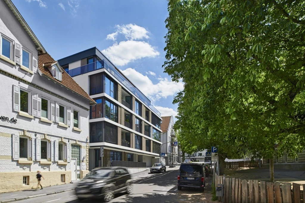 Projekte An einer sonnigen Straße in Schorndorf steht ein modernes Glasgebäude neben älteren, traditionellen Häusern; dort parken und fahren Autos, und grüne Bäume spenden Schatten.