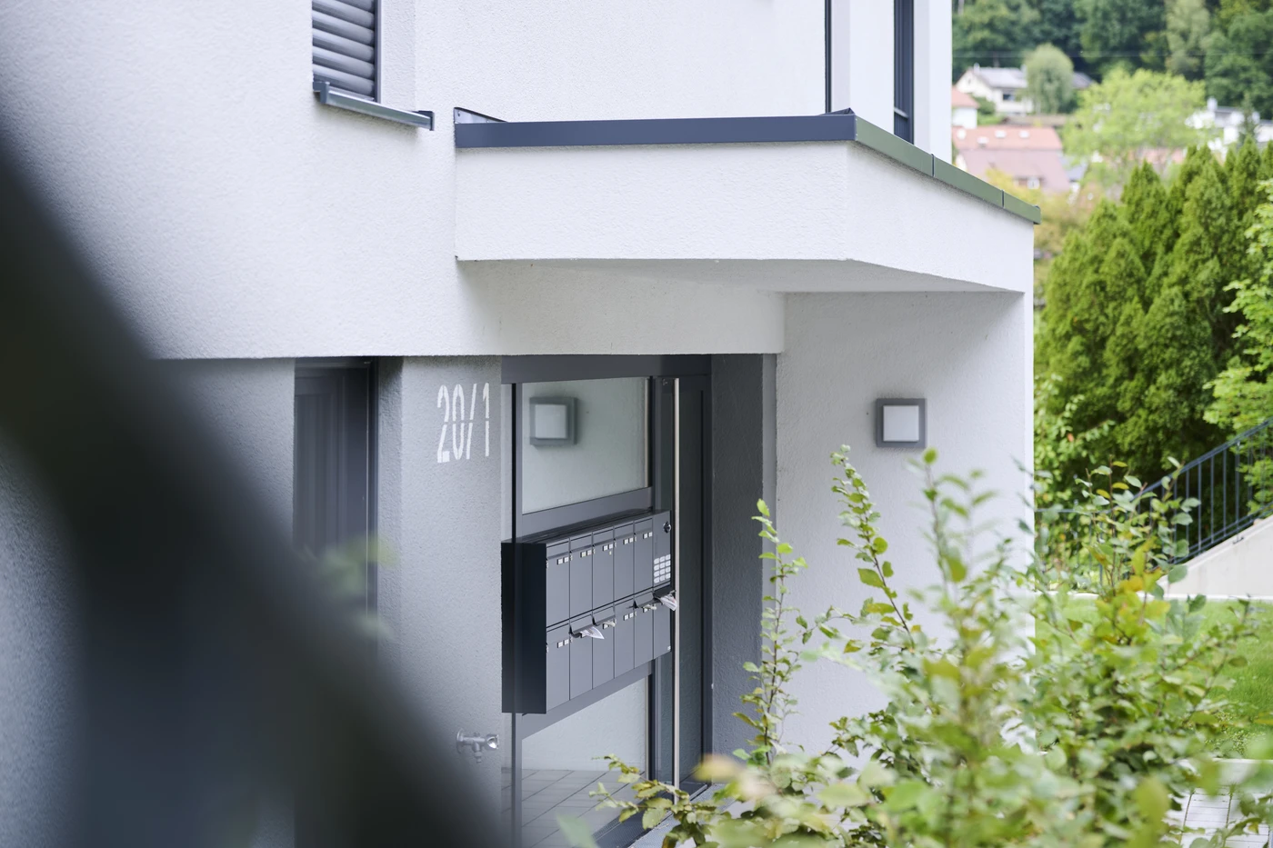 Projekte Eingang eines modernen, weißen Mehrfamilienhauses mit durchdachtem Design, mit mehreren an der Wand angebrachten Briefkästen, üppiger Bepflanzung im Vordergrund und einem Teilblick auf nahegelegene Häuser in Schorndorf im Hintergrund.