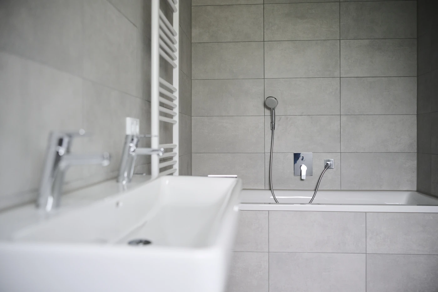 Projekte Modernes Badezimmer mit grau gefliesten Wänden, einem weißen Waschbecken mit zwei Wasserhähnen, einer weißen Badewanne, einer Handbrause und einem Handtuchheizkörper im Leitergestalt. Die Gestaltung wirkt klar und minimalistisch – ganz im Sinne des durchdachten Entwurfs und der raffinierten Ästhetik, die für SCHATZ Architektur charakteristisch sind.