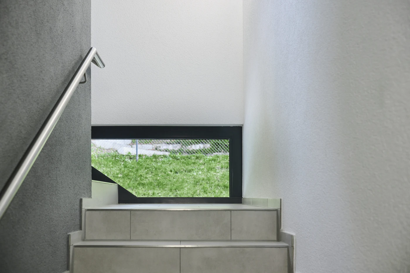 Eine Treppe mit hellen Fliesen und einem Metallhandlauf führt hinauf zu einem Fenster mit Blick auf grünes Gras und einen Zaun, was den durchdachten Entwurf widerspiegelt, der für die moderne Architektur in Schorndorf charakteristisch ist. Weiße und graue Wände säumen die Treppe und unterstreichen die sorgfältige Gesamtplanung des gesamten Raums.