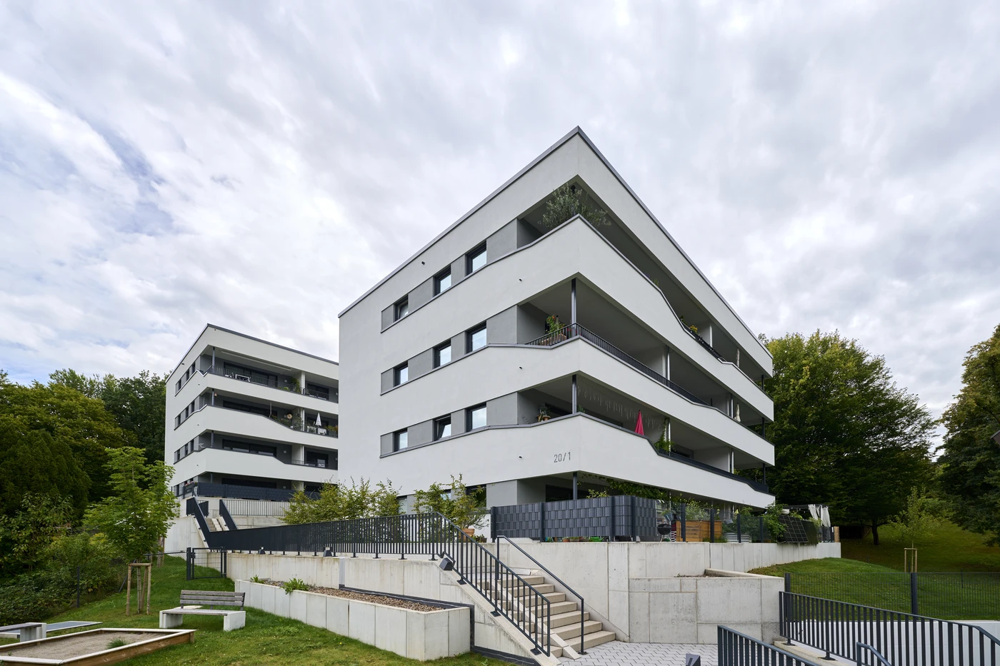 Modernes weißes Mehrfamilienhaus mit zahlreichen Balkonen, umgeben von Grünflächen und Bäumen, unter bewölktem Himmel. Treppen und eine Rampe führen zum Eingang und zeugen von durchdachtem Entwurf und sorgfältiger Projektentwicklung in jeder Bauphase.