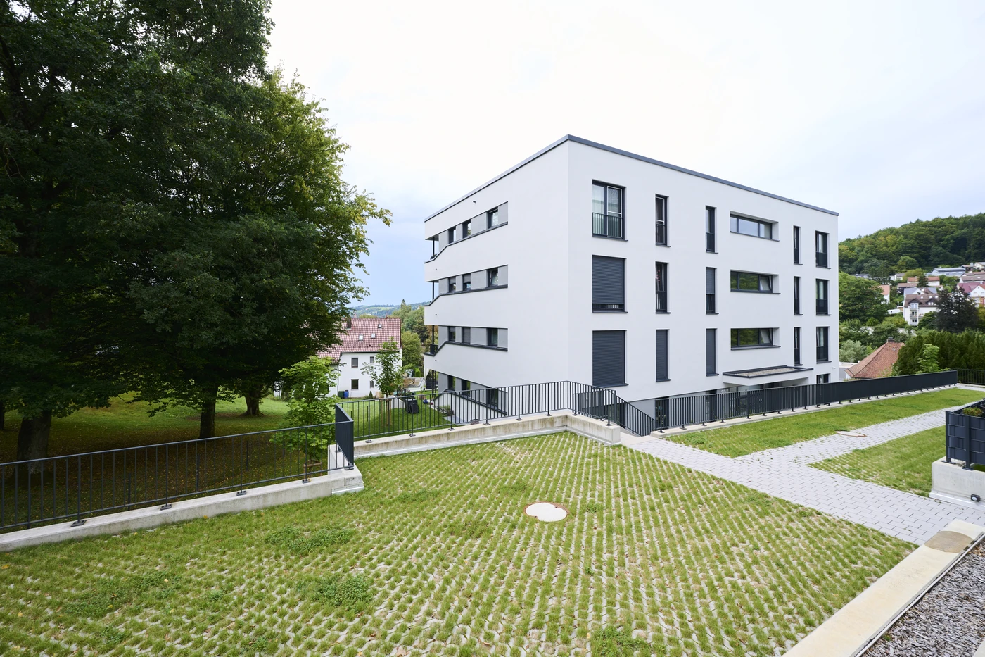 Ein modernes, weißes Mehrfamilienhaus mit vielen Fenstern und dunklen Fensterläden, umgeben von grünen Bäumen, Rasenflächen und einem gepflasterten Gehweg, auf einem leicht abfallenden Gelände in einer vorstädtischen Umgebung – ein vorbildliches Projekt von SCHATZ Architektur, das die Kompetenz des Büros in der Generalplanung über alle Leistungsphasen hinweg unter Beweis stellt.