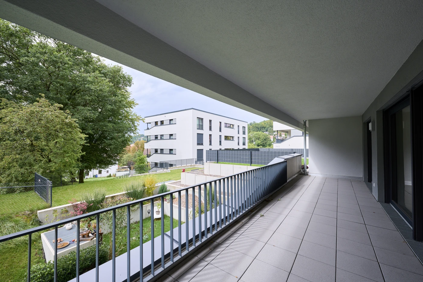 Ein großzügiger, überdachter Balkon mit Fliesenboden bietet an einem bewölkten Tag den Blick auf eine grüne Rasenfläche, Bäume und ein modernes, weißes Wohnhaus im Hintergrund – ein idealer Aussichtspunkt, um die zeitgenössische Generalplanung in Schorndorf zu bewundern.