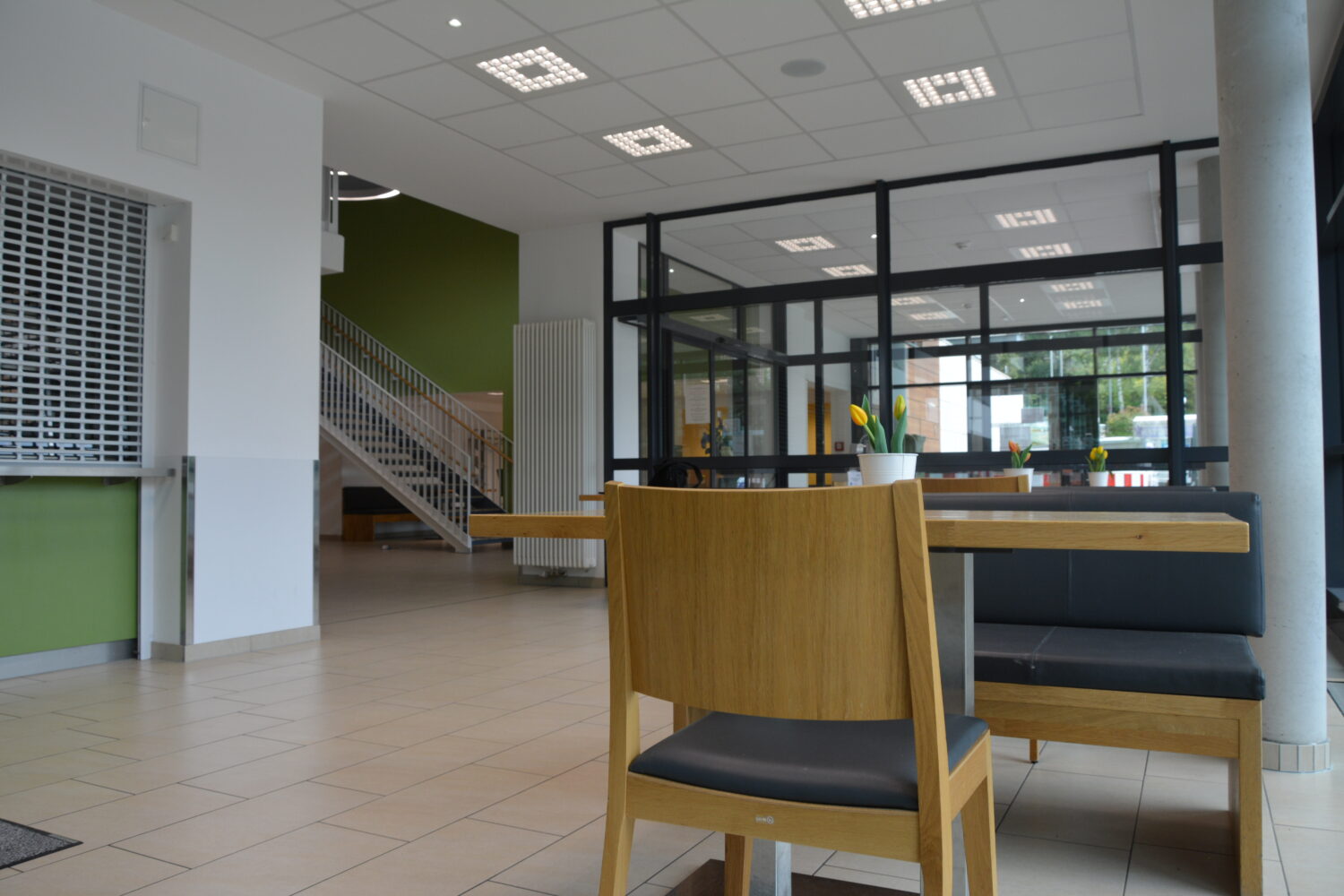 Ein modernes Cafeteria-Interieur mit Holzstühlen und einer Sitzbank, im Hintergrund ist eine Treppe zu sehen, die in ein Obergeschoss führt.